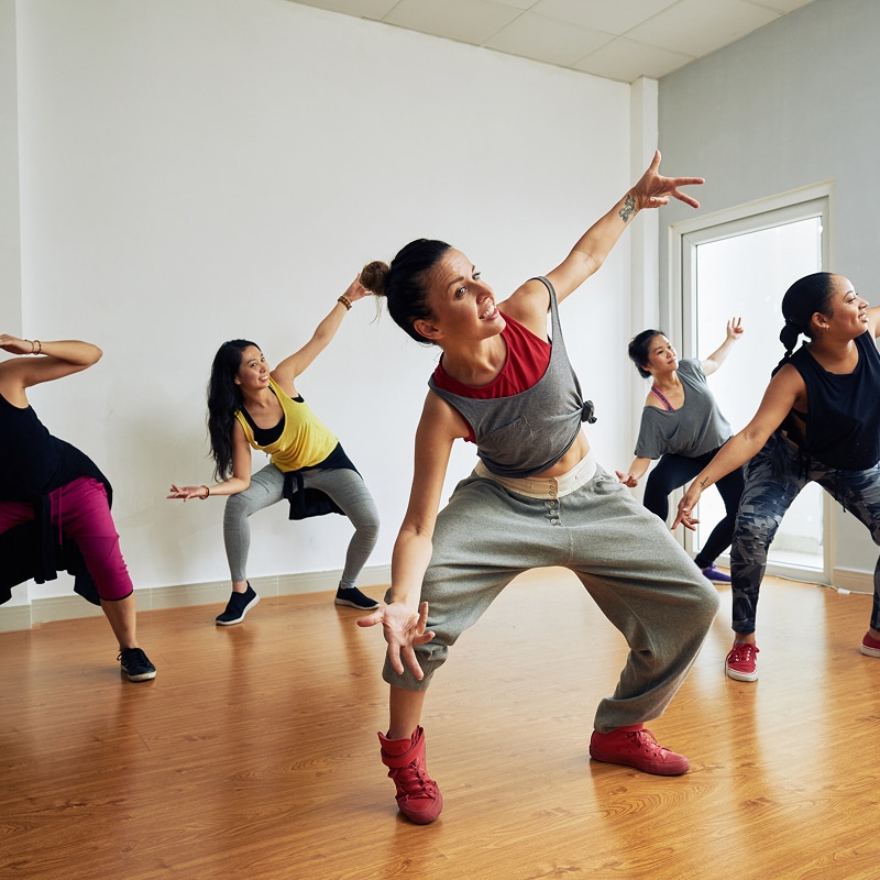 Cinq femmes dansent avec énergie dans un studio. La danseuse au premier plan, souriante, est en pleine action, les bras levés.