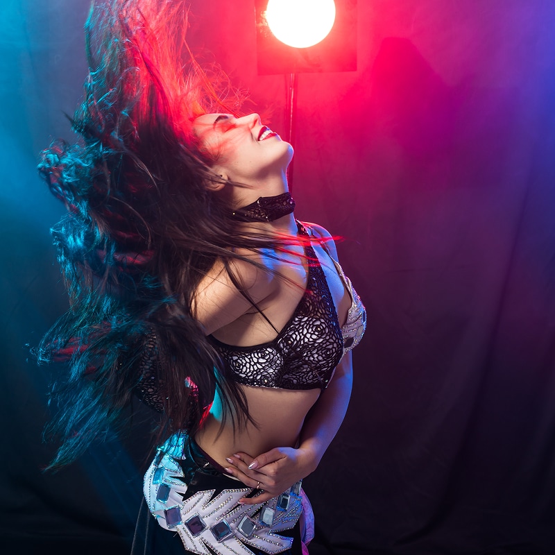 Jeune danseuse souriante, cheveux flottants, en costume de scène noir et argenté, sous lumières rouge et bleue.