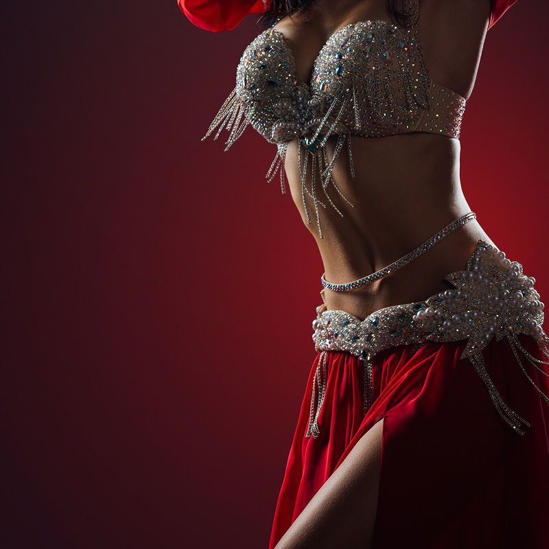 Danseuse orientale élégante en costume rouge étincelant, parée de bijoux argentés et perles, sur fond dégradé bordeaux.
