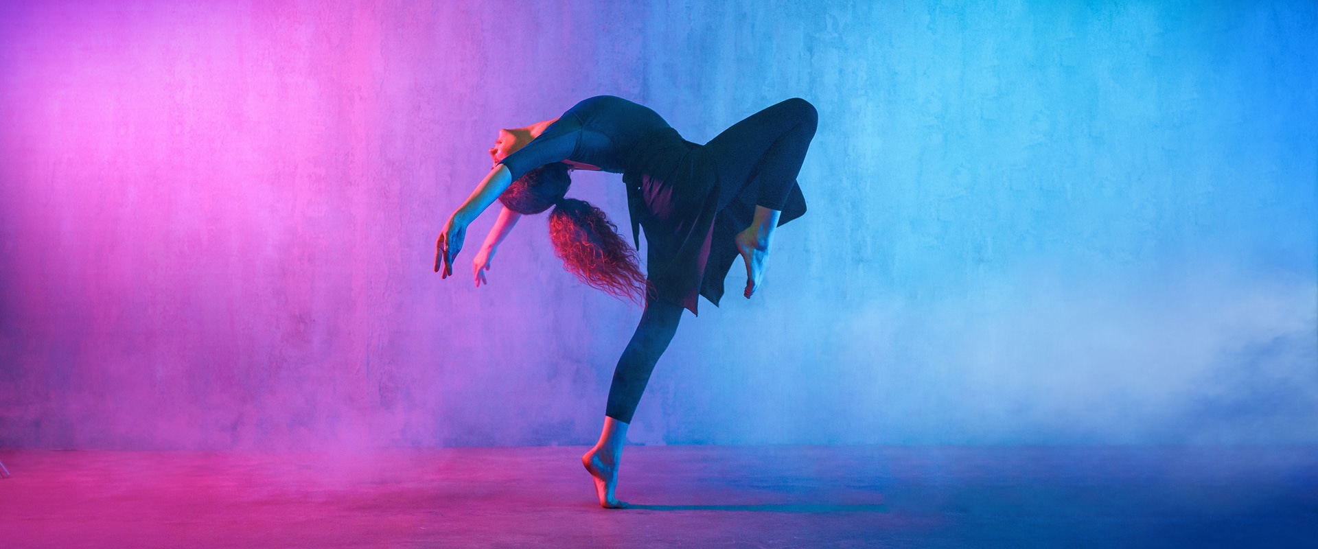 Danseuse en pleine extension arrière, vêtue de noir, sur un fond de studio dégradé rose et bleu néon, avec une atmosphère vaporeuse.