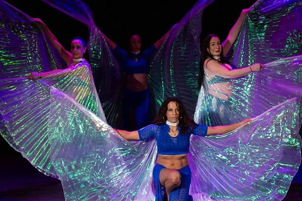 Cinq danseuses en costumes bleus avec des ailes iridescentes vertes et violettes, effectuant une chorégraphie dynamique sur scène.