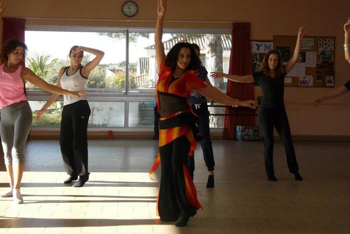 Cinq femmes en pleine danse dans un studio lumineux. Une danseuse au centre porte un costume orange vif, bras levé. Les autres adoptent des poses de danse.