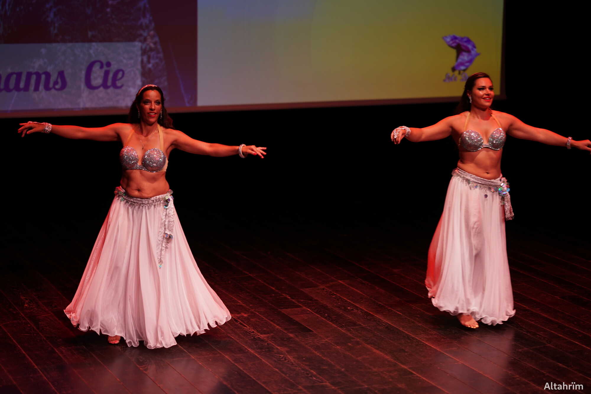 Spectacle de Danse Orientale : Deux Danseuses sur Scène Deux femmes souriantes en costumes de danse orientale (soutien-gorge argenté, jupe blanche) sur scène, bras étendus. Écran avec "ams Cie".
