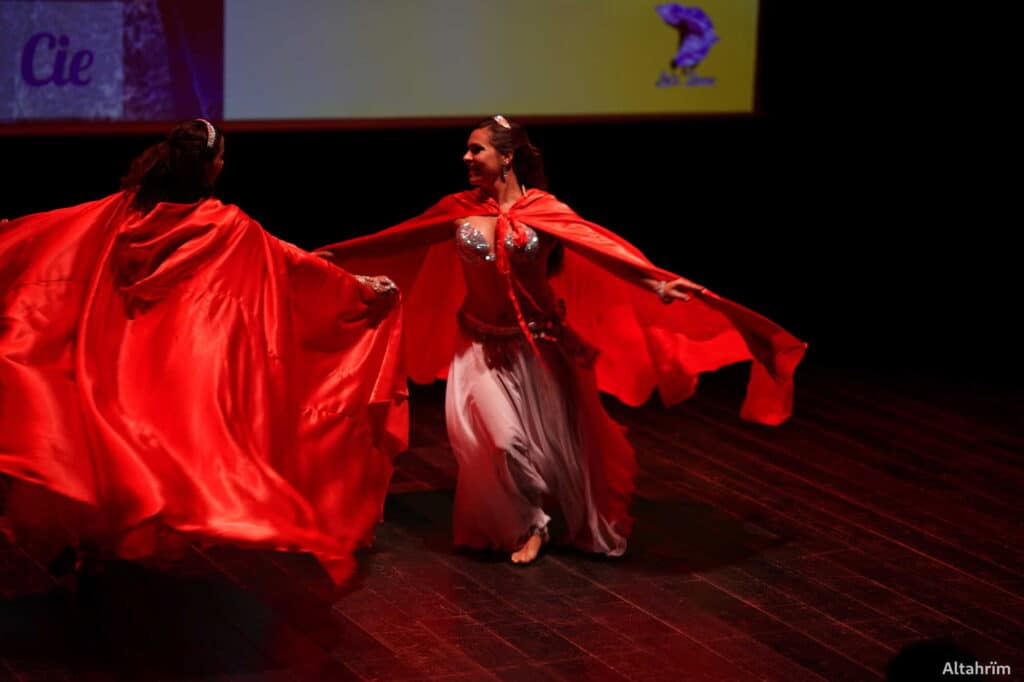 Deux danseuses souriantes en capes rouges flottantes et costumes de danse orientale sur scène en bois. Fond écran "Cie".