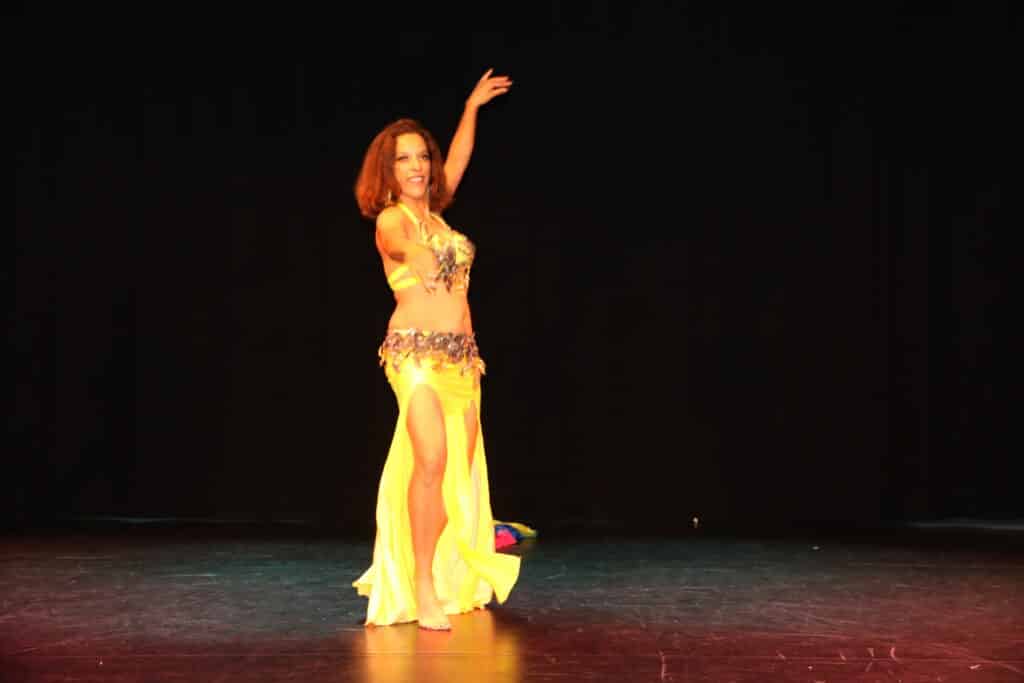 Danseuse orientale en costume jaune: Spectacle captivant Danseuse du ventre souriante en costume jaune brillant sur scène sombre, bras levé, captivant le public avec sa performance.
